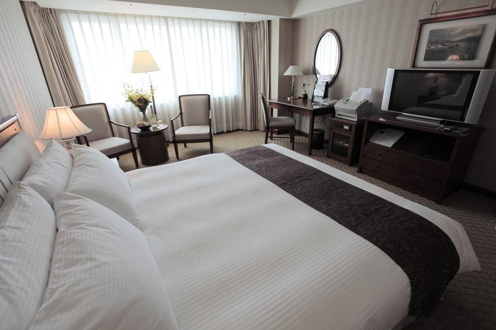 Evergreen Laurel Hotel - Taichung Extérieur photo The photo shows a well-furnished hotel room. In the foreground, there is a large bed with white bedding and a dark-colored throw at the foot. Beside the bed, there is a bedside lamp. 

In the background, natural light filters through large windows co
