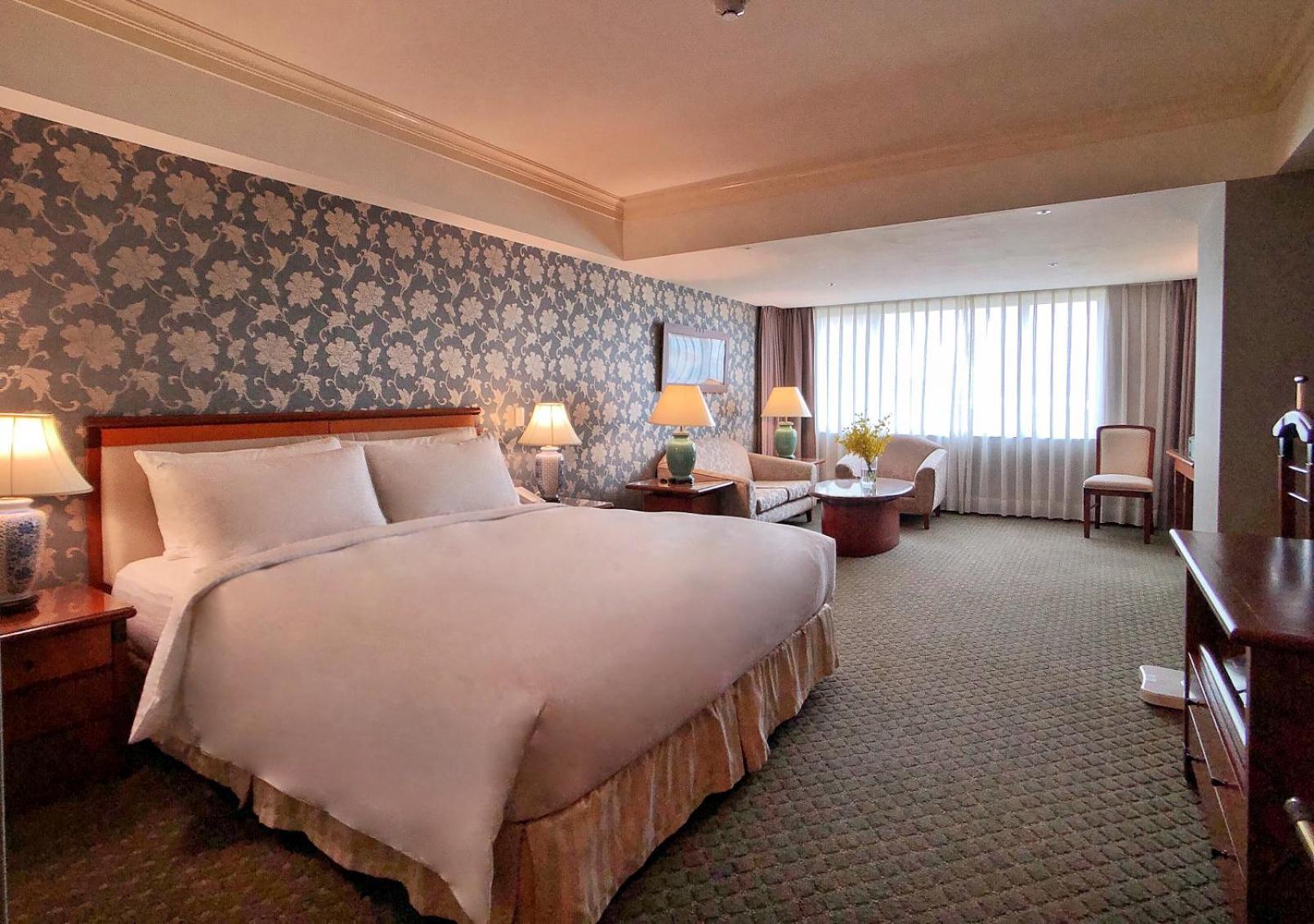 Evergreen Laurel Hotel - Taichung Extérieur photo The photo shows a hotel room with a spacious and comfortable layout. It features a large bed with white bedding, placed against a decorative wallpapered wall. On either side of the bed, there are lamps providing soft lighting. In addition to the bed,