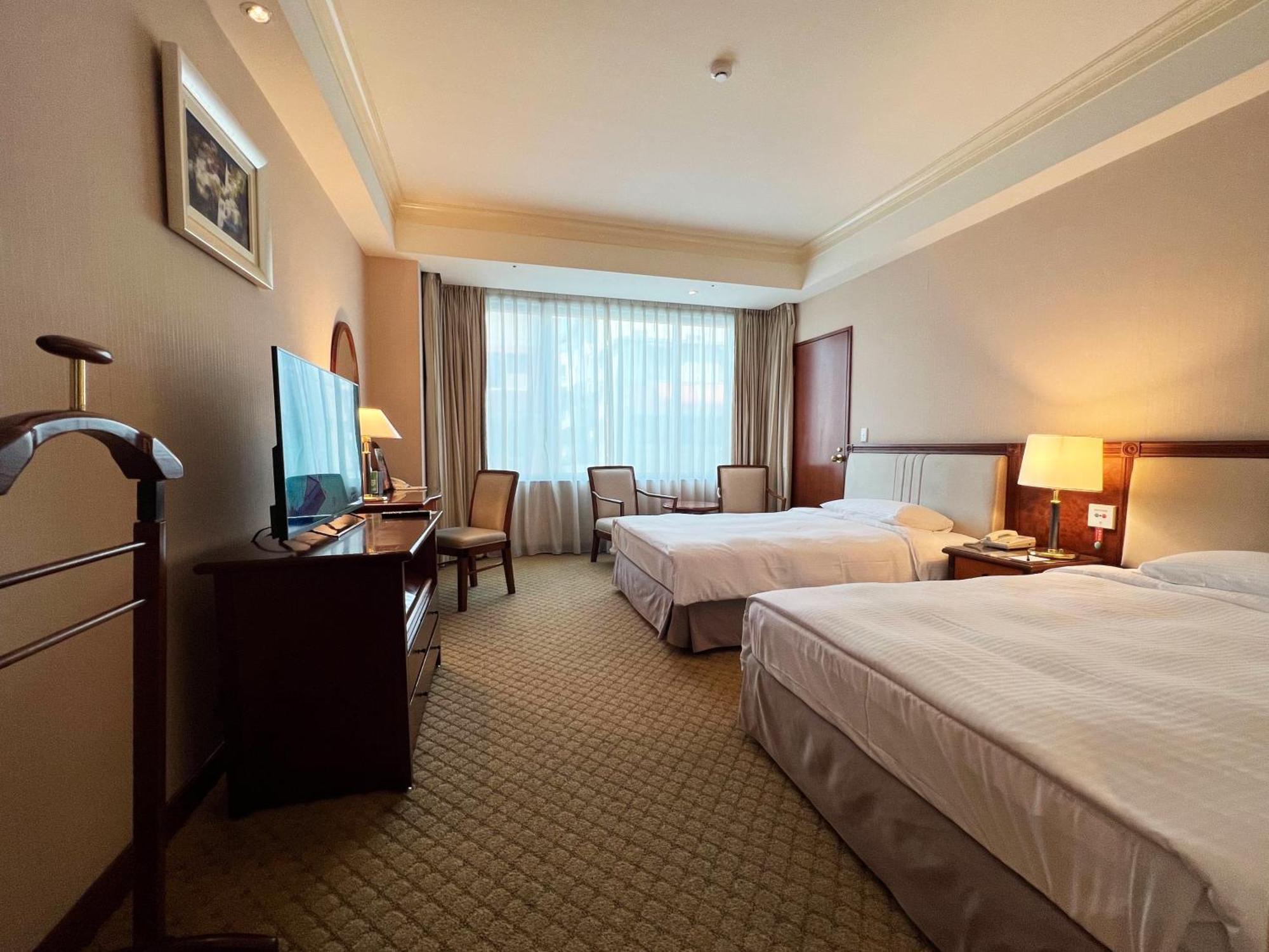 Evergreen Laurel Hotel - Taichung Extérieur photo The photo shows a hotel room featuring two twin beds placed parallel to each other. The room has a warm color palette and includes a large window with sheer curtains allowing natural light to illuminate the space. There is a wooden desk and two chair