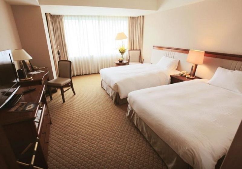 Evergreen Laurel Hotel - Taichung Extérieur photo The photo shows a hotel room featuring two twin beds with white bedding. There's a chair positioned near one of the beds and a desk with a computer setup against one wall. Large windows allow natural light to enter the room, and light-colored curtain