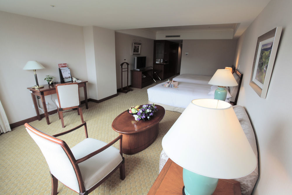 Evergreen Laurel Hotel - Taichung Extérieur photo The photo shows an interior view of a hotel room. It features two beds, a coffee table with a small decorative arrangement of flowers, and a comfortable chair. There's a desk and a lamp, along with another larger lamp on the table. In the background,