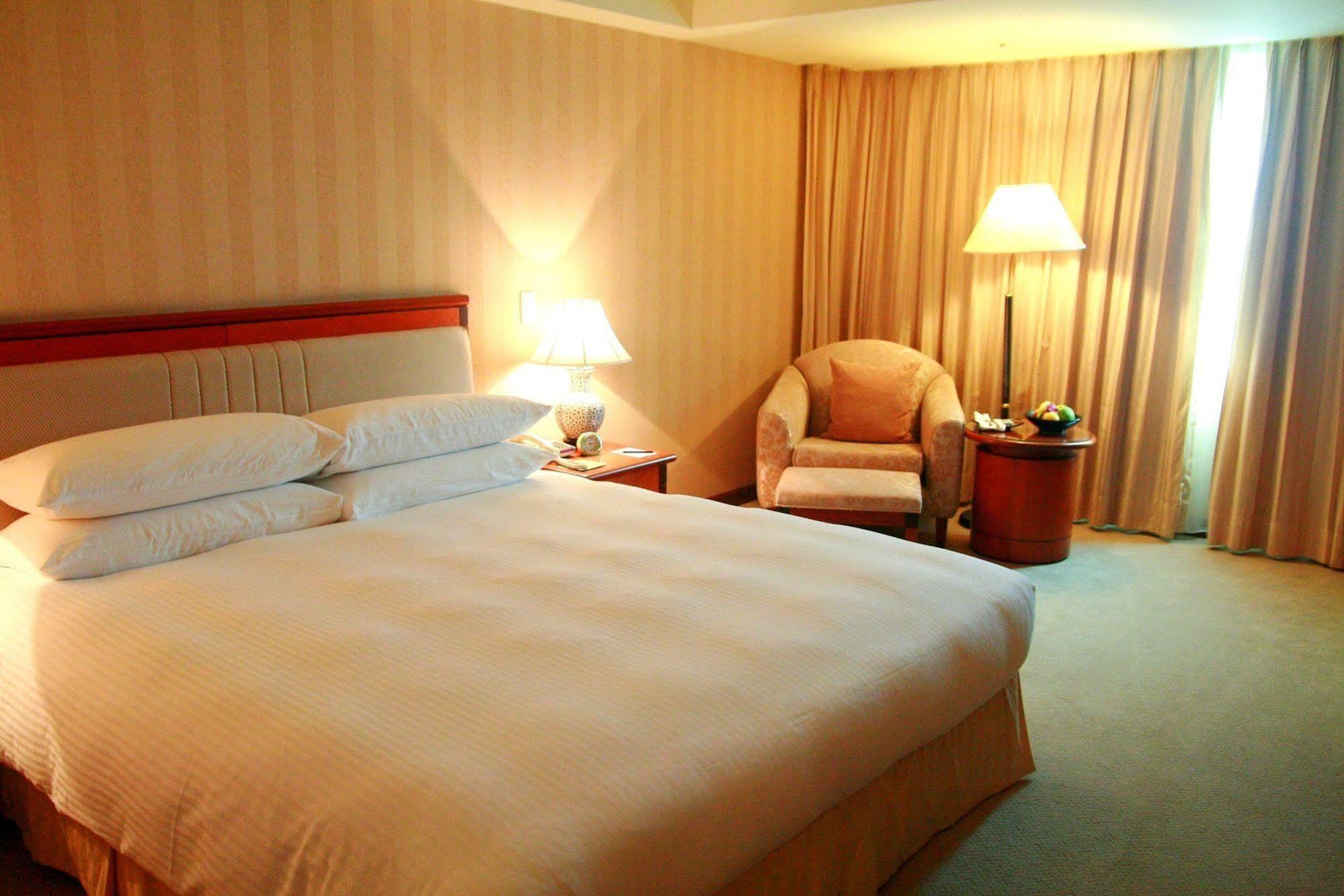 Evergreen Laurel Hotel - Taichung Extérieur photo The photo shows a hotel room containing a large bed with white linens, flanked by two bedside lamps. To one side, there is a cozy armchair and a small side table with a fruit bowl or snacks. The walls are decorated in a soft, neutral color, and there