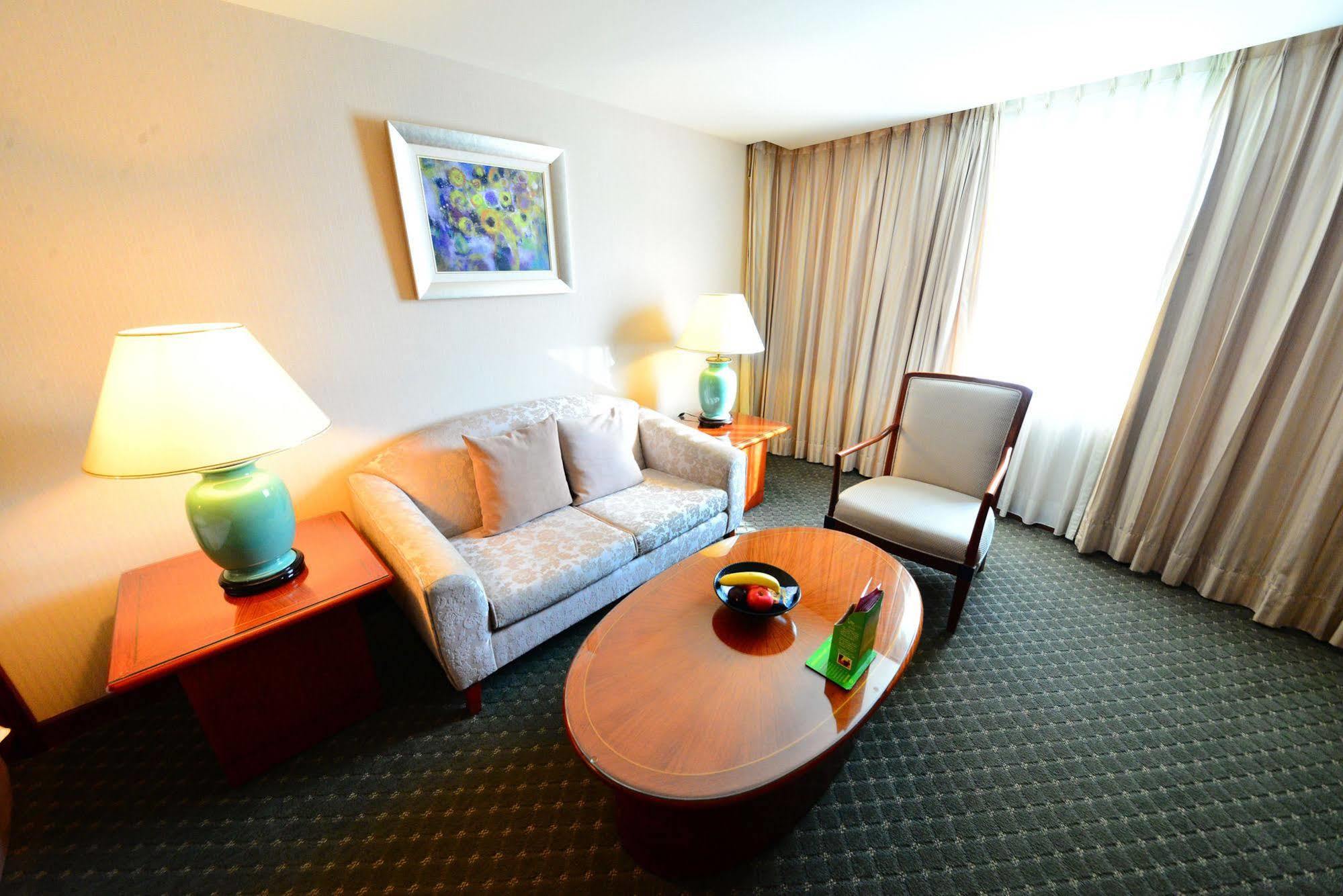 Evergreen Laurel Hotel - Taichung Extérieur photo The photo shows a cozy living space, likely a hotel room or a lounge area. It features a light-colored sofa with cushions, a wooden coffee table in the center, and a comfortable armchair beside it. There are two lamps with turquoise bases on side tab