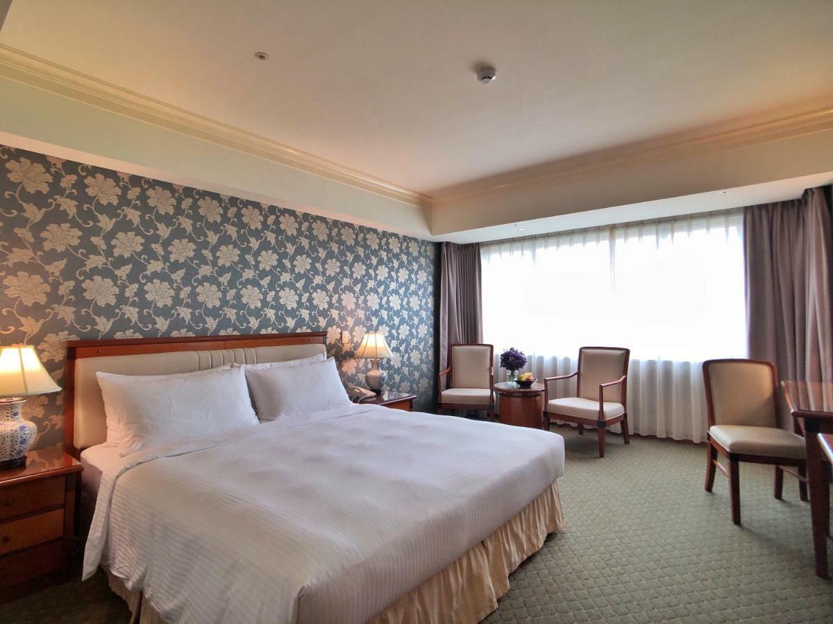 Evergreen Laurel Hotel - Taichung Extérieur photo The photo shows a hotel room that features a large bed with a white bedspread positioned against a decorative floral wallpaper. There are two bedside lamps on wooden nightstands flanking the bed. To the side of the room, there are two chairs and a sm