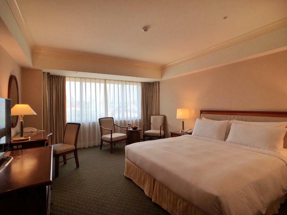 Evergreen Laurel Hotel - Taichung Extérieur photo The photo shows a hotel room featuring a large bed with white linens, a wooden bedside table with a lamp, and a flat-screen television. There are two chairs with a small table next to them positioned by a large window, which has sheer curtains lettin