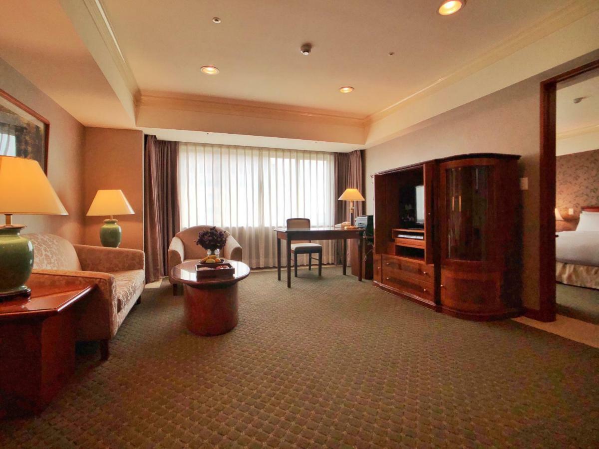 Evergreen Laurel Hotel - Taichung Extérieur photo The photo shows a cozy hotel suite or living area. There is a sofa and an armchair arranged around a small coffee table. A desk with a chair is positioned near a large window covered with sheer curtains that allow natural light to enter the room. Two