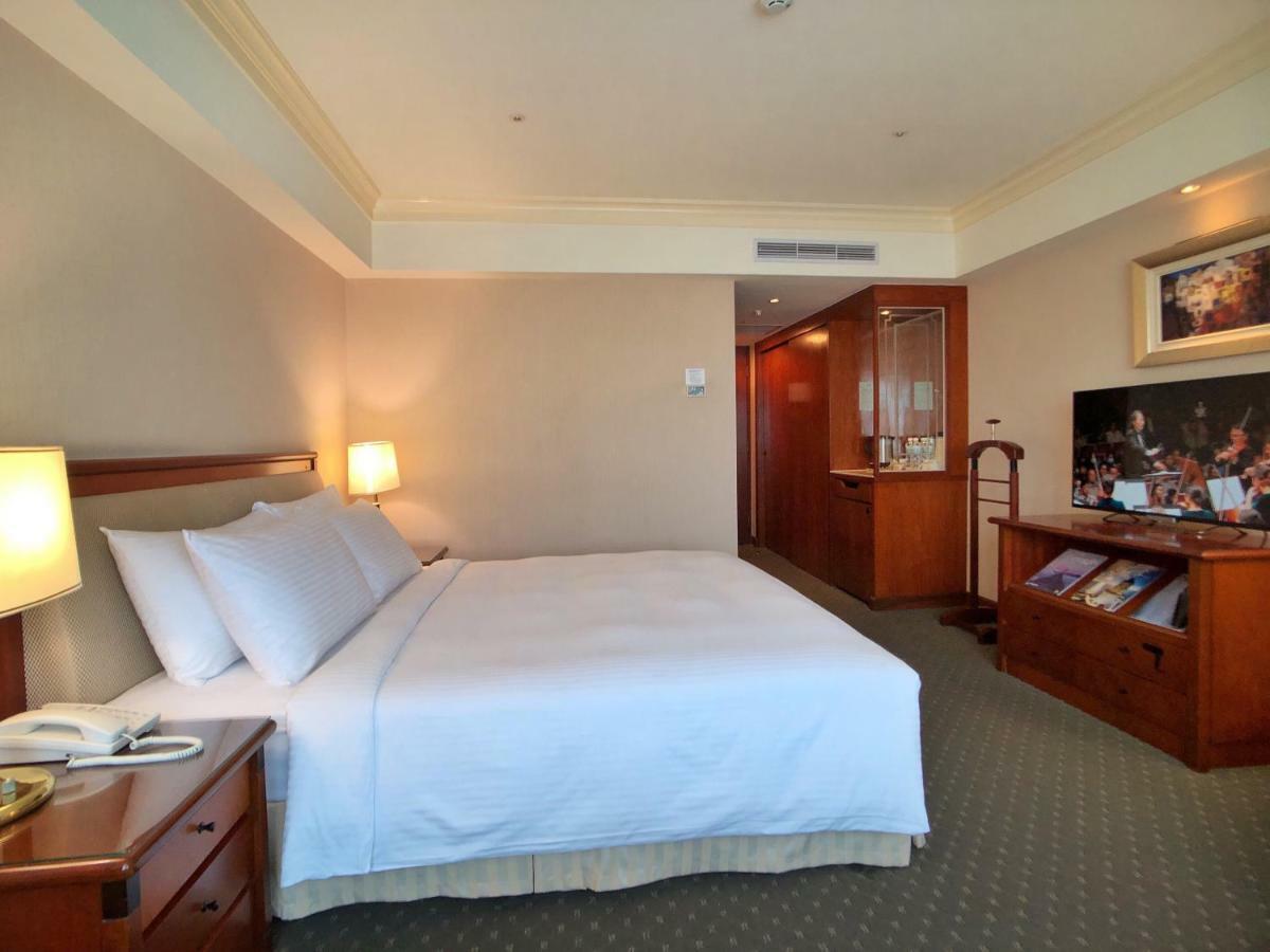 Evergreen Laurel Hotel - Taichung Extérieur photo The photo shows a hotel room featuring a comfortable bed with white linens and pillows, positioned against a light-colored wall. There is a wooden bedside table with a lamp and an open book next to the bed. On the opposite side, there’s a large flat-