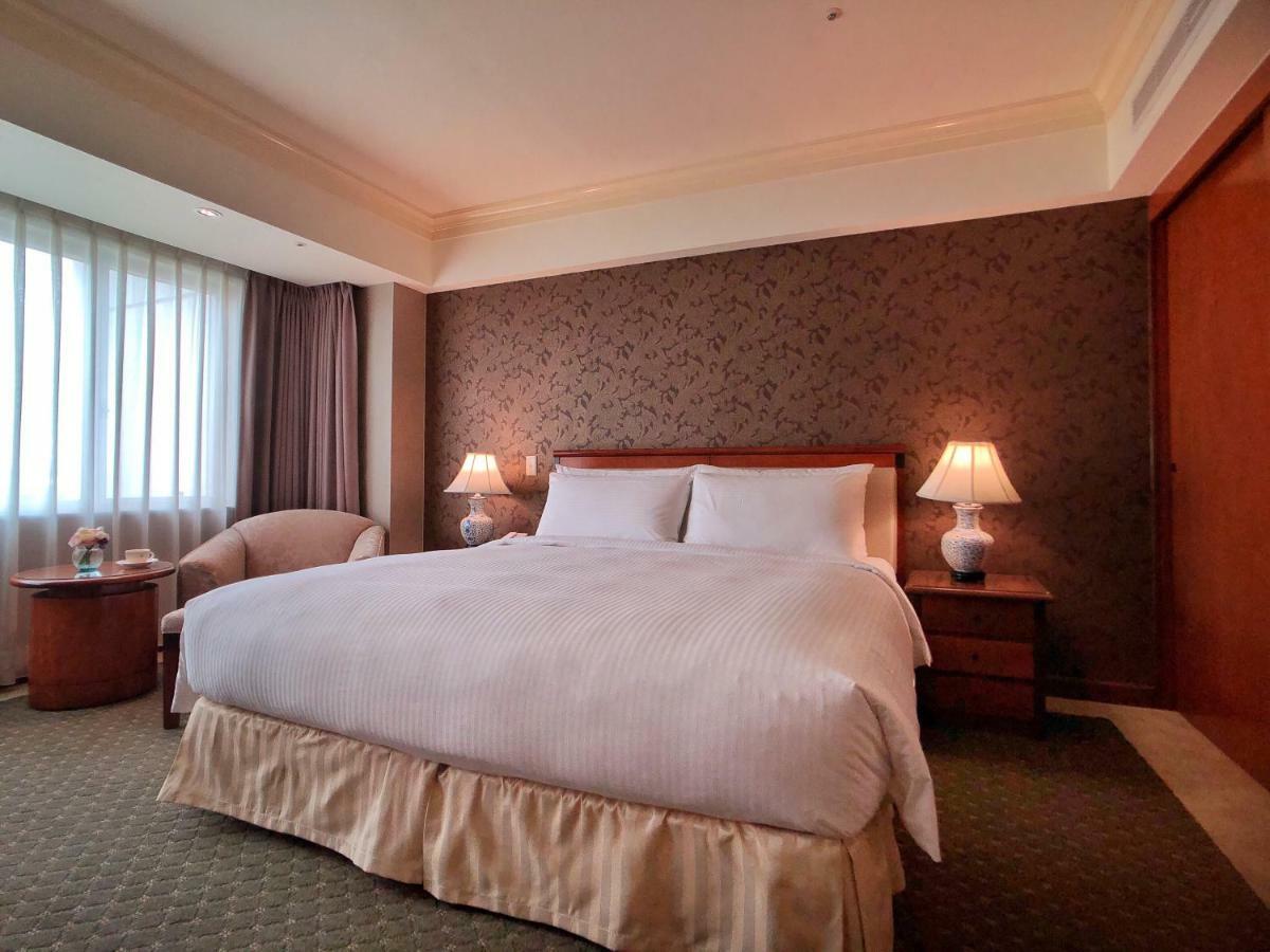 Evergreen Laurel Hotel - Taichung Extérieur photo The photo shows a neatly arranged hotel room featuring a large bed with white linens and a light-colored bed skirt. On either side of the bed, there are small wooden nightstands with lamps. To the left of the bed, there's a comfortable chair and a ro