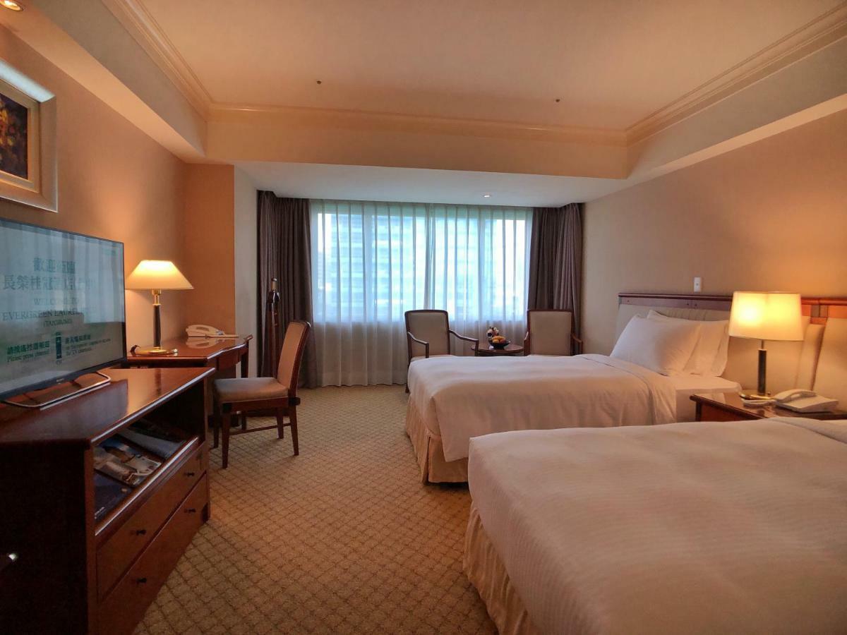 Evergreen Laurel Hotel - Taichung Extérieur photo The photo shows a hotel room featuring two twin beds with white bedding. There's a television mounted on a wooden dresser, and next to it, a comfortable chair and a small table are positioned. The room has large windows that allow natural light to en