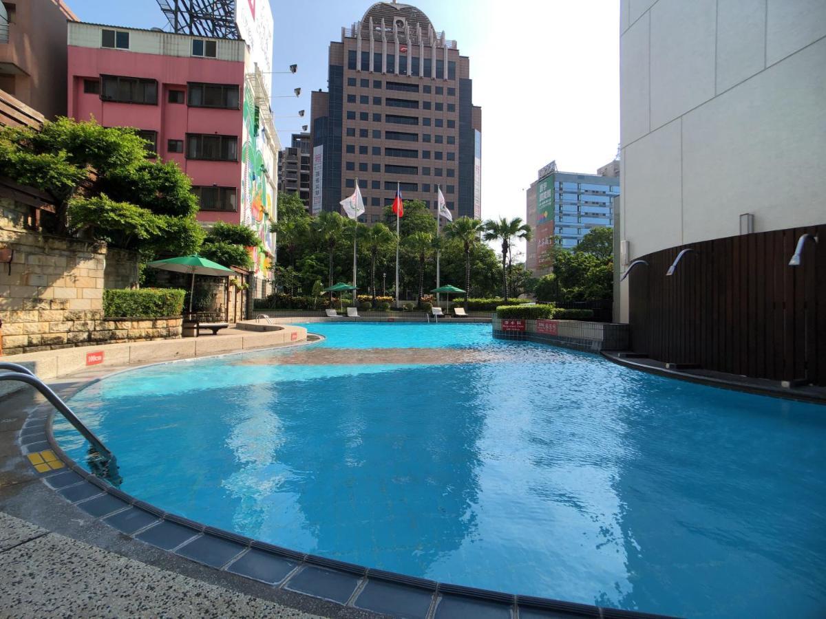Evergreen Laurel Hotel - Taichung Extérieur photo The photo shows a swimming pool area set in an outdoor environment. The pool has a clear blue water and a gently curving shape. Surrounding the pool are some green trees and plants, along with a few umbrellas providing shade. In the background, there